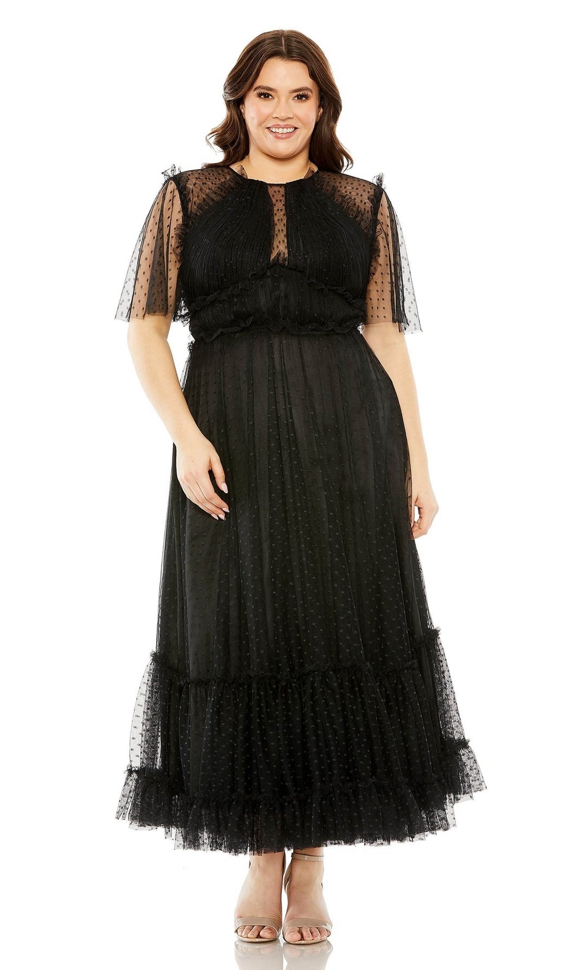 A woman stands and smiles wearing the Long Plus-Size Prom Dress 68568 by Mac Duggal, a high-neck, black ankle-length gown featuring a sheer polka-dot overlay and ruffled details.